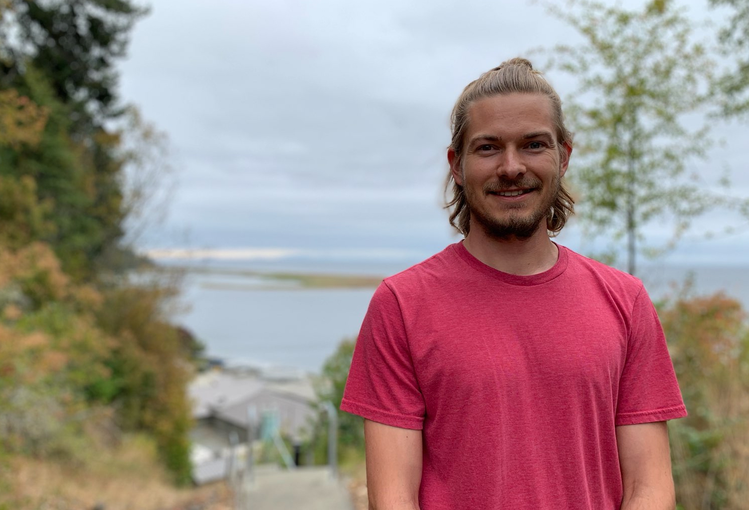 Image of a white man with long hair and a beard in a red tshirt standing in front of a scenic coastline