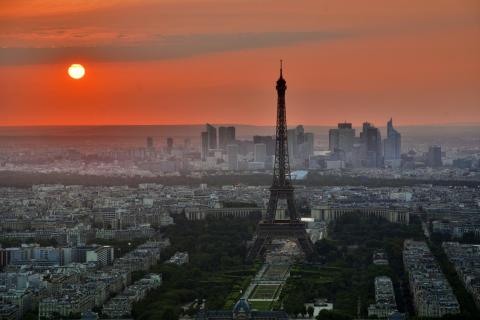 The glowing orange Sun shines over a lightly hazy Paris, the Eifel Tower standing erect, just right of center-frame.