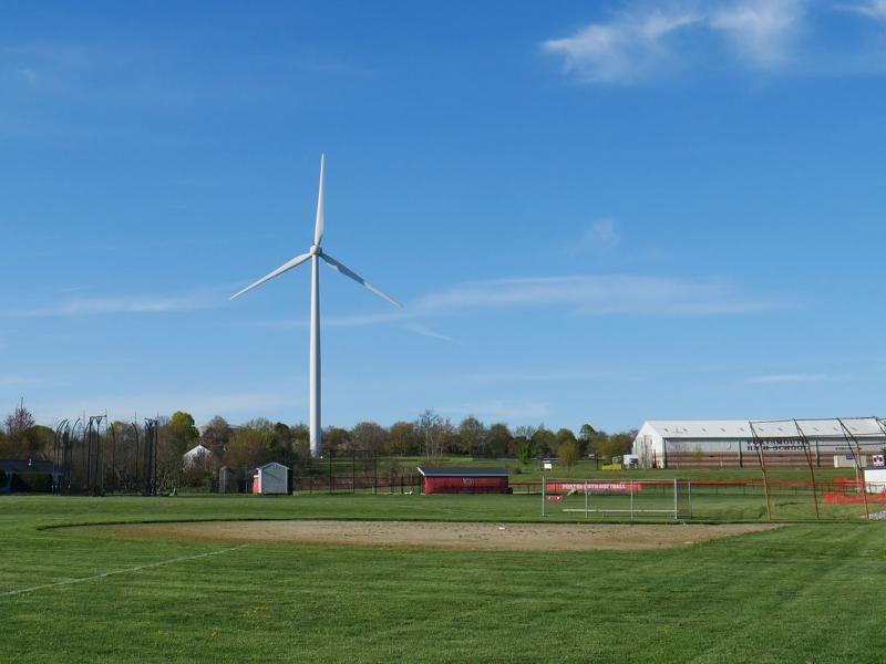 A wind turbine near a small baseball field. There are also white buildings to the right of the turbine.